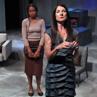 A woman with dark hair and a brown shirt standing behind a woman in a blue dress